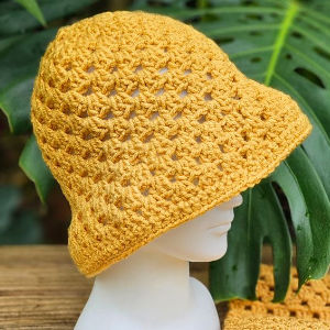 Un chapeau jaune au crochet est présenté sur la tête d'un mannequin blanc, avec une grande feuille verte en arrière-plan. Le chapeau présente un motif ouvert en dentelle et un large bord.