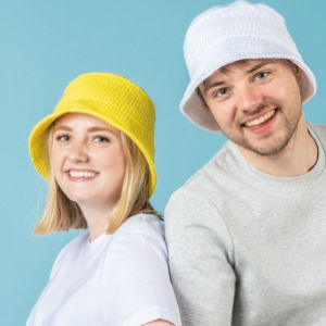 Une femme souriante portant un chapeau jaune et un homme portant un chapeau blanc posent ensemble sur un fond bleu clair.