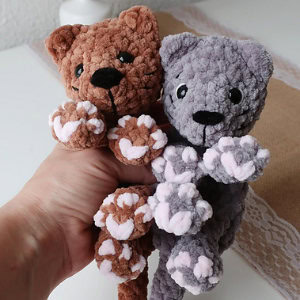 Une main tient deux chats en peluche, l'un brun et l'autre gris, à la fourrure douce et aux coussinets roses et blancs, face à l'appareil photo sur un fond clair.