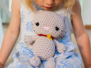 Un jeune enfant vêtu d'une robe à fleurs bleu clair et blanche tient sur ses genoux un chat au crochet - un chat en peluche crocheté avec un collier rouge et une étiquette jaune.