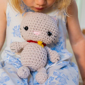 Une jeune enfant en robe blanche et bleue tient un chat au crochet, une poupée de chat gris avec des yeux noirs, un nez rose et un collier rouge avec un nœud jaune.