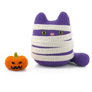 Un chat au crochet violet, enveloppé dans des bandages de momie blancs, est assis à côté d'une petite citrouille orange crochetée avec une tige verte et un visage de lanterne noire.