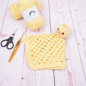 Couverture en crochet jaune avec une tête de canard, à côté d'un fil jaune, d'un crochet orange et de ciseaux noirs sur une surface en bois clair.