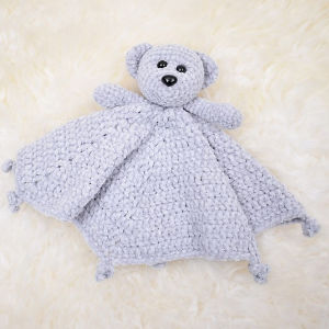 Un tendre ourson crocheté gris clair avec une tête d'ours et des bras attachés à un petit corps ressemblant à une couverture, sur un fond crème duveteux.