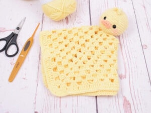 Un doudou plat au crochet jaune avec une tête de canard est posé sur une surface en bois blanc, à côté d'une paire de ciseaux, d'un crochet et d'une pelote de laine jaune.