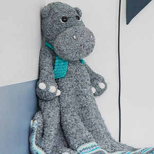 Un hippopotame gris en peluche avec une écharpe bleue et blanche est assis debout contre un mur. Son corps s'étend vers l'extérieur, ressemblant à une couverture aux bords bleus et blancs.