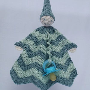 Une poupée crochetée avec un corps en couverture verte et bleue à motifs en zigzag, un chapeau pointu et une tétine attachée au centre. La poupée a une tête et des mains simples et rondes.