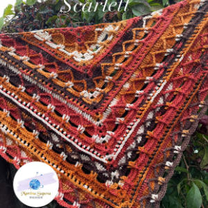 Un châle triangulaire coloré en crochet avec des motifs complexes dans les tons rouge, orange, marron et crème est drapé à l'extérieur. Un petit logo circulaire avec du texte se trouve dans le coin inférieur gauche.
