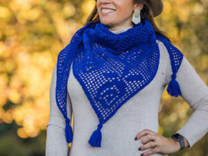Une femme portant un châle triangulaire bleu vif, crocheté avec des pompons - fabriqué à partir d'un modèle chèche gratuit - se tient à l'extérieur, souriante. L'arrière-plan est estompé par des feuillages jaunes et verts.