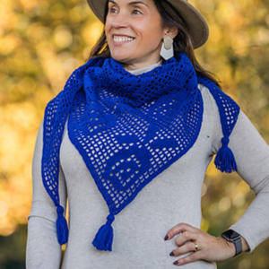 Une femme portant un chapeau et une robe beige sourit tout en modelant un châle triangulaire crocheté bleu vif avec des pompons. L'arrière-plan est brouillé par des feuillages d'automne.