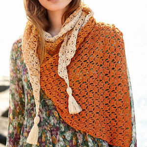 Une personne portant une robe à fleurs et un châle crocheté orange et crème à glands drapé sur ses épaules. Le châle est orné d'un motif de dentelle détaillé et de deux glands bien visibles.