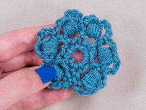 Une main avec du vernis à ongles bleu tient une fleur bleue crochetée avec des noppes au crochet en rond au centre et des boucles en forme de pétales, sur un fond beige clair.
