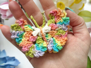 Une main tient un papillon coloré au crochet, réalisé avec des fils multicolores jaunes, verts, bleus, orange, roses et violets. Son corps blanc et ses antennes vertes se détachent sur des fleurs floues en arrière-plan.