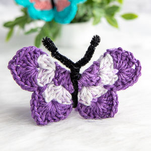 Un papillon fait main au crochet, de couleur violette et blanche, avec un corps en cure-pipe noir, est présenté sur une surface blanche, avec une plante floue en arrière-plan.
