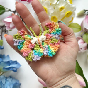 Une main tient un petit papillon coloré au crochet dans des tons pastel, entouré de fleurs artificielles en arrière-plan.
