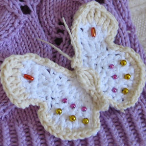Un papillon crocheté aux ailes crème et blanches, décoré de petites perles rouges, jaunes et roses, est présenté sur un tricot texturé violet.
