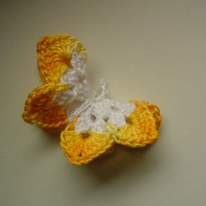 Un petit papillon au crochet, blanc au centre et aux ailes jaunes et orange, posé sur une surface unie de couleur claire.