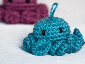 Une petite pieuvre bleue en crochet porte clé avec des yeux noirs est posée sur une surface blanche, avec une pieuvre violette en crochet floue en arrière-plan.