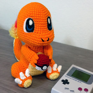 Une peluche Charmander crochetée tient une petite Poké Ball, assise sur une table à côté d'une Nintendo Game Boy grise classique.
