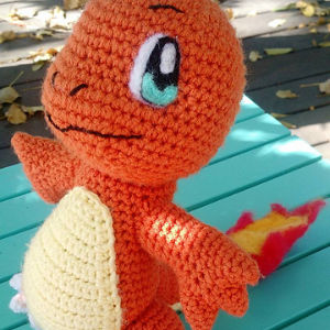 Une salamèche au crochet, peluche crochetée ressemblant au Pokémon orange au ventre crème et à la queue enflammée, est posée sur une surface turquoise avec des feuilles en arrière-plan.