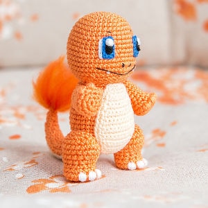 Un dinosaure orange crocheté avec de grands yeux bleus, un ventre crème, de petites griffes blanches et une queue orange duveteuse, debout sur une surface beige avec des motifs orange et blancs.
