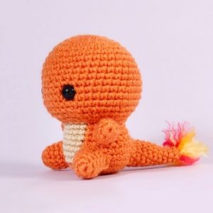 Une peluche dinosaure orange crochetée avec un ventre crème, de grands yeux noirs et un bout de queue en fil jaune et rouge, sur un fond violet clair.