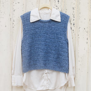 Une chemise blanche à col large est superposée à un gilet en tricot bleu texturé sans manches, tous deux suspendus à un cintre contre un mur blanc cassé et abîmé.