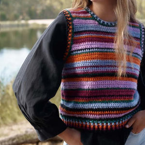 Une personne portant un gilet coloré, rayé et crocheté par-dessus une chemise noire à manches longues se tient à l'extérieur, près d'un lac calme avec des arbres en arrière-plan.