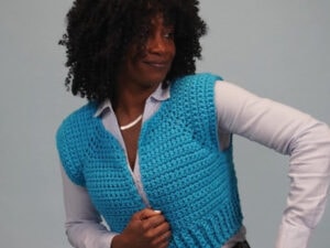 Une femme aux cheveux bouclés porte un gilet bleu vif au crochet, inspiré par un tuto de pull sans manche femme au crochet, sur une chemise à boutons bleu clair, debout sur un fond uni et regardant sur le côté avec un léger sourire.
