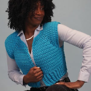 Une femme aux cheveux bouclés portant une chemise bleu clair et un gilet crocheté bleu vif pose sur un fond clair uni.