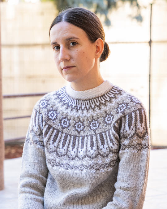 Une personne aux cheveux bruns raides tirés vers l'arrière porte un pull en maille à motifs et est assise à l'extérieur. Elle a une expression neutre et regarde légèrement sur le côté. L'arrière-plan est flou.