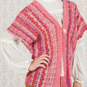 Une personne porte un gilet en crochet rose et rouge à rayures verticales sur un chemisier blanc texturé à manches longues. Le cardigan comporte un seul bouton et est ouvert sur le devant. L'arrière-plan est un papier peint à motifs.