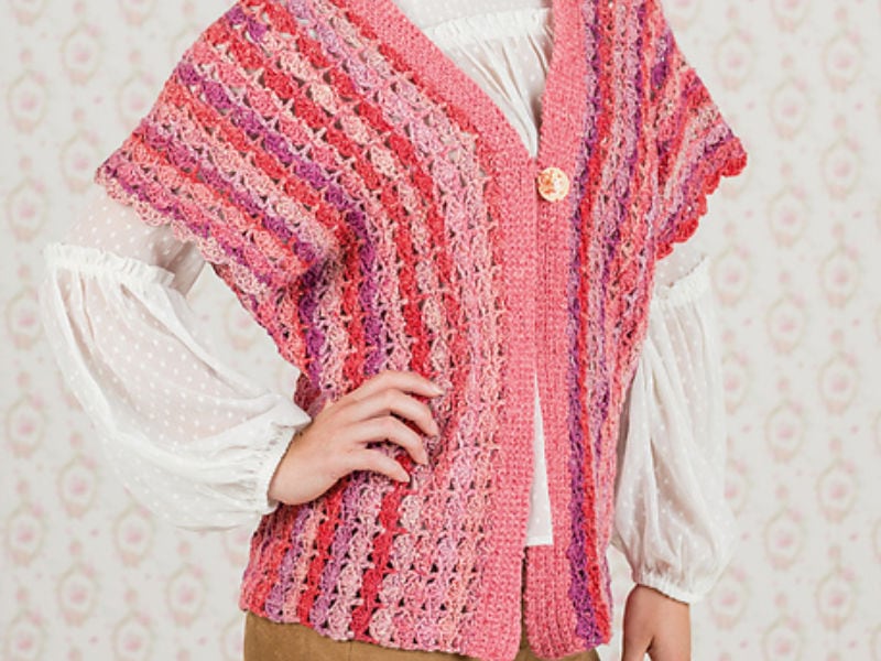 Une personne porte un gilet sans manche au crochet texturé et multicolore, rose et rouge, sur un chemisier blanc à manches bouffantes et un pantalon beige, sur un fond à motifs clairs.
