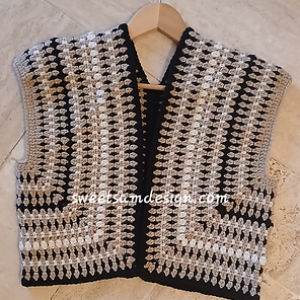 Un gilet crocheté à motif géométrique noir, blanc et gris est présenté sur un cintre en bois sur un fond clair. Le motif présente des rangées de points texturés et un devant ouvert.