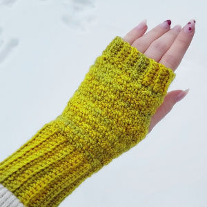Une main portant un gant sans doigts crocheté jaune-vert vif est étendue sur un fond enneigé. Le gant couvre le poignet et la paume, laissant les doigts exposés. Les ongles sont manucurés avec des pointes rouges.