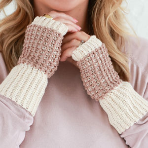 Une personne portant un haut mauve et des gants tricotés sans doigts de couleur beige et marron clair tient ses mains près de son visage, montrant le design texturé des gants.