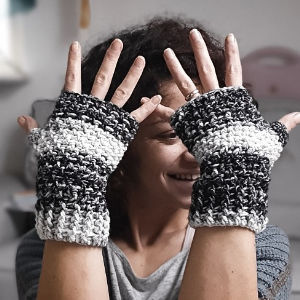 Une femme sourit et lève les mains pour montrer de gros gants sans doigts en crochet à rayures noires et blanches.