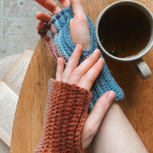 Une personne portant des gants tricotés sans doigts tient une autre main gantée, avec une tasse de thé et un livre ouvert sur une table en bois à proximité.