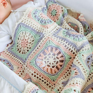 Un bébé fait la sieste sous une couverture crochetée aux couleurs pastel, avec des motifs carrés complexes dans les tons rose, vert, bleu et crème.