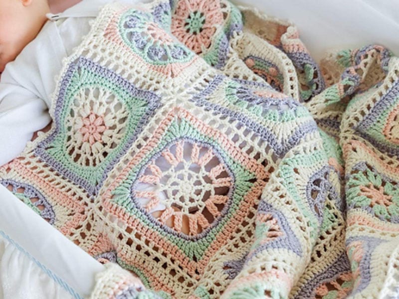 Un bébé dort couvert d'un modèle de crochet en coton aux couleurs pastel, avec des motifs carrés complexes dans des tons de rose, de bleu, de vert et de crème.