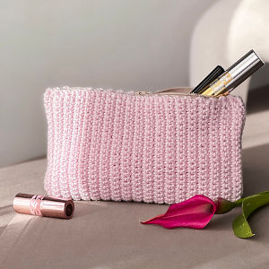Une trousse de toilette rose pâle en tricot est posée sur une surface avec des articles de maquillage à l'intérieur, un rouge à lèvres rose et une fleur de calla magenta placés à proximité.