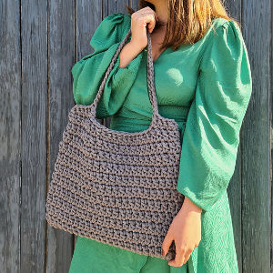 Femme en robe verte tenant à deux mains un grand sac au crochet gris, debout devant une clôture en bois.