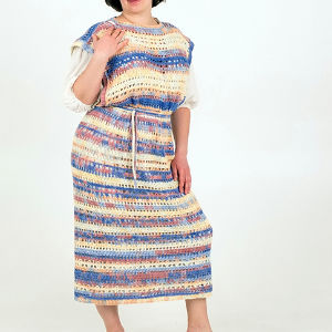 Une personne pose dans une robe en crochet multicolore, faite à la main, avec une ceinture à la taille. La robe comporte des rayures bleues, jaunes, blanches et orange et est portée sur un haut blanc. L'arrière-plan est uni et de couleur claire.
