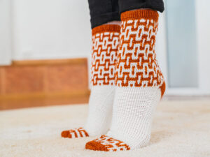 Une personne portant des chaussettes tricotées ornées d'un motif « mosaïque au crochet » orange et blanc se tient debout sur un tapis de couleur claire. Les chaussettes ont la pointe et le bord-côte orange, avec un motif répétitif sur la partie supérieure.