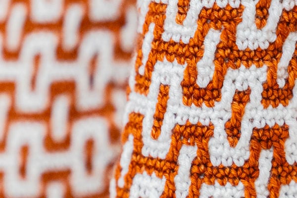 Gros plan sur un tissu tricoté orange et blanc, orné d'un motif géométrique en forme de labyrinthe inspiré de la « mosaïque au crochet ». Le motif se compose de formes entrelacées qui mettent en valeur les détails texturés du fil.