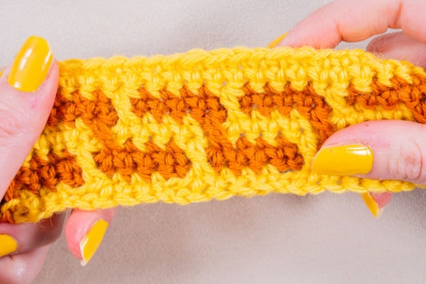 Gros plan sur des mains aux ongles vernis en jaune, tenant un morceau de tissu au crochet réalisé avec du fil jaune et orange, présentant un motif en mosaïque au crochet en zigzag.