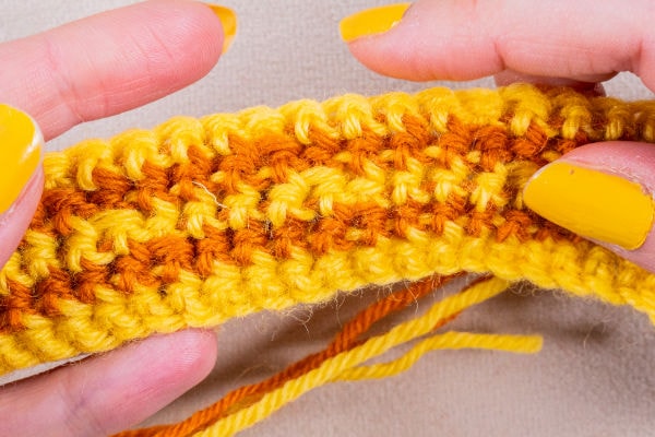 Gros plan sur des mains aux ongles vernis en jaune tenant une création « mosaïque au crochet » aux couleurs vives, réalisée à partir de fils jaunes et orange.