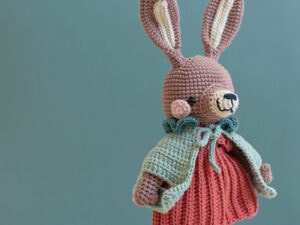 Une peluche en crochet avec de longues oreilles, un nez rose et un visage souriant porte une robe rouge et une cape verte en tricot, posée sur un fond uni sarcelle.