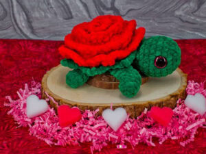 Une tortue au crochet avec un corps vert et une coquille rouge ressemblant à une rose est posée sur une plaque de bois, entourée de papier déchiqueté rose et de décorations en forme de cœur sur une surface rouge à motifs.