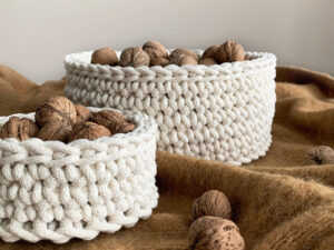Deux gros paniers blancs crochetés à la main avec de la grosse laine, inspirés du tuto au crochet, sont remplis de noix non écalées sur une surface brune texturée. Les noix éparpillées renforcent l'impression de confort et de rusticité de la scène.